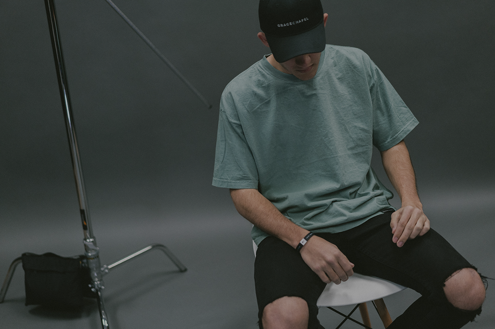 A man in a casual pose, wearing a soft mint green t-shirt and black cap with the text 'Apparel Boss,' seated on a white stool. This image captures a behind-the-scenes moment, highlighting the relaxed yet stylish appeal of Apparel Boss's custom apparel in a professional photoshoot setting.